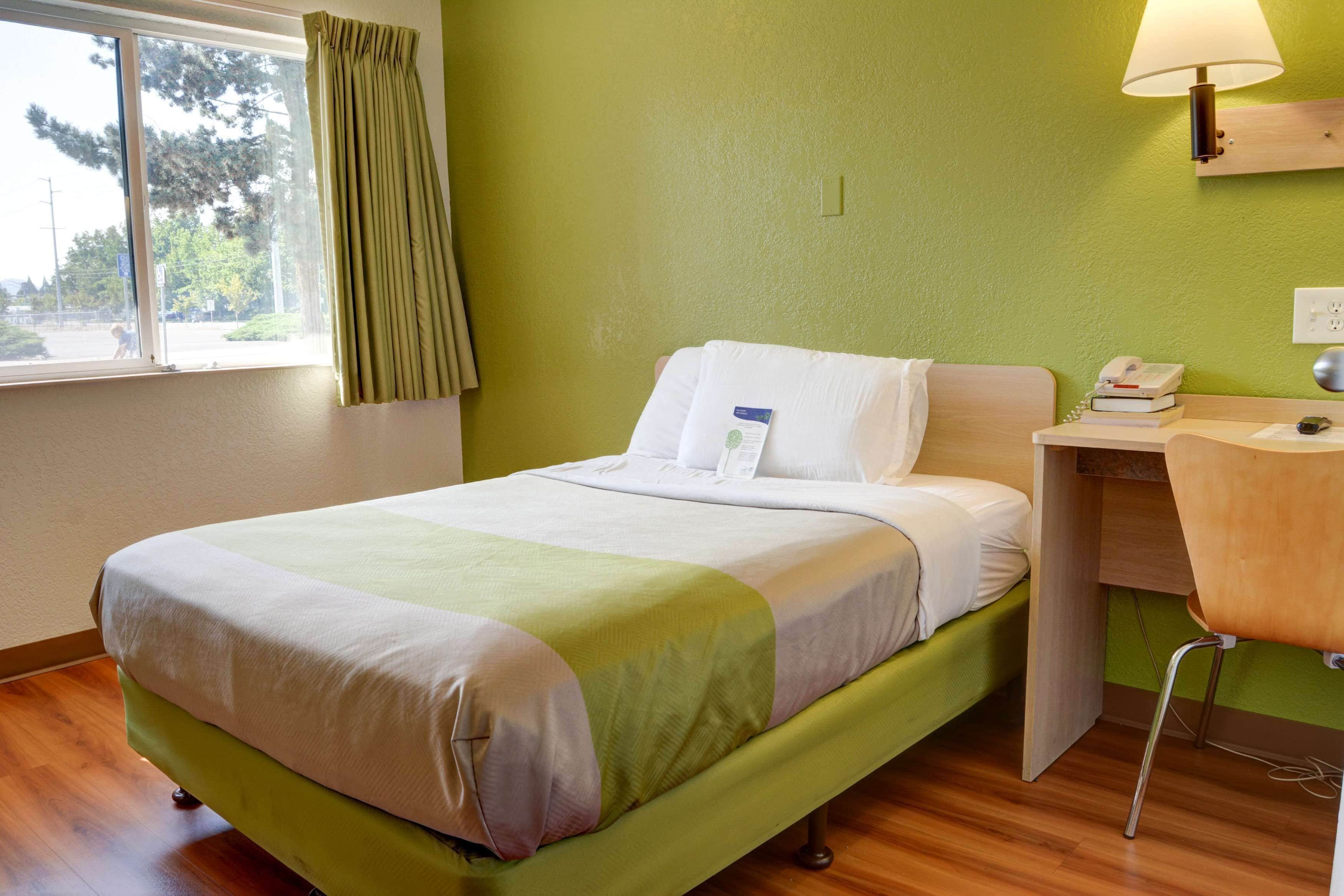 Une chambre d’hôtel avec un lit, un bureau, une chaise, une lampe et une fenêtre avec des rideaux.