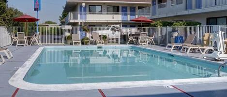 Piscina retangular com espreguiçadeiras e guarda-sóis, ladeada por uma cerca branca, com um edifício ao fundo.