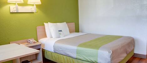 Quarto de hotel com cama, mesa de cabeceira, luminária e pequena escrivaninha com cadeira.