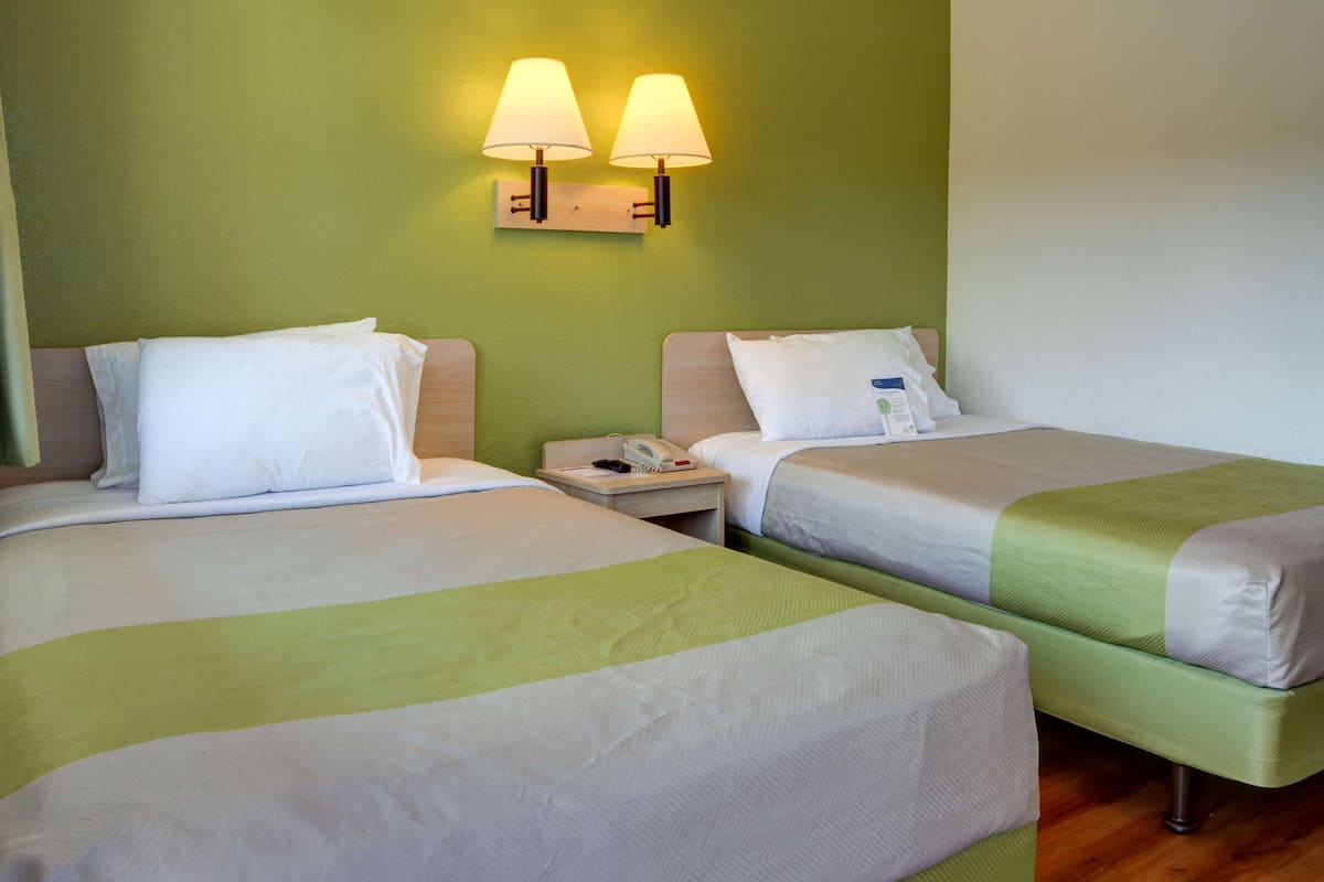 Quarto de hotel com duas camas, cada uma com lençóis brancos e detalhes em verde. Há duas luminárias fixadas na parede acima das camas, e uma mesa de cabeceira com um telefone e uma garrafa.