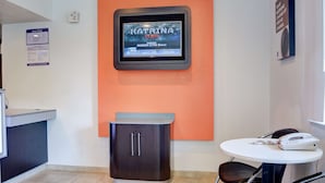 Un área de recepción con una pantalla digital mostrando precios de habitaciones, un mueble pequeño, una mesa con un teléfono y un letrero en la pared.
