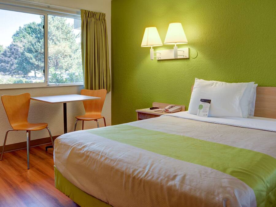 A hotel room with a bed, two chairs, a table, a nightstand with a phone, and a window with curtains.