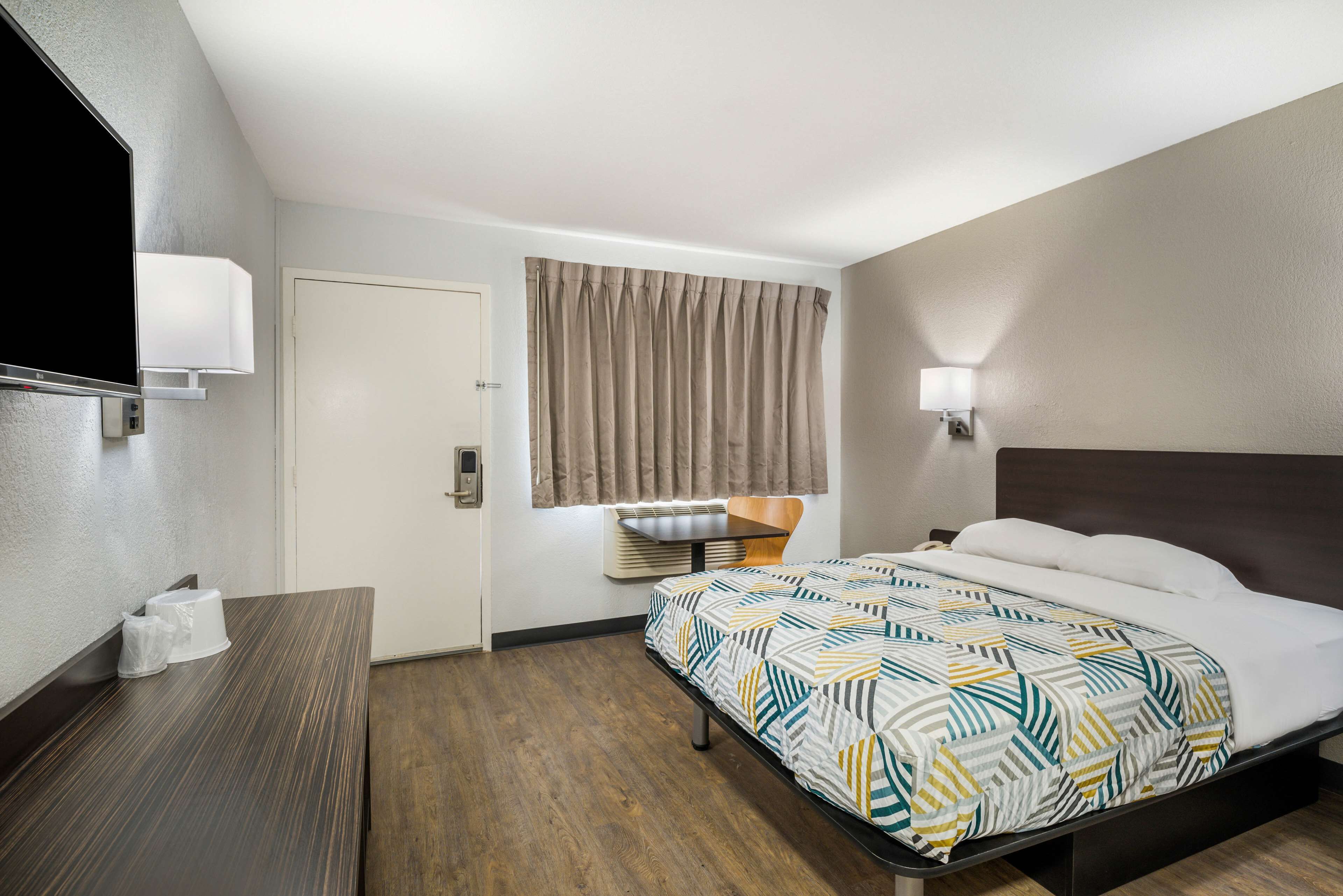 Ein Hotelzimmer mit Bett, Schreibtisch, Fernseher, einem Fenster mit Vorhängen und einer Wandlampe.