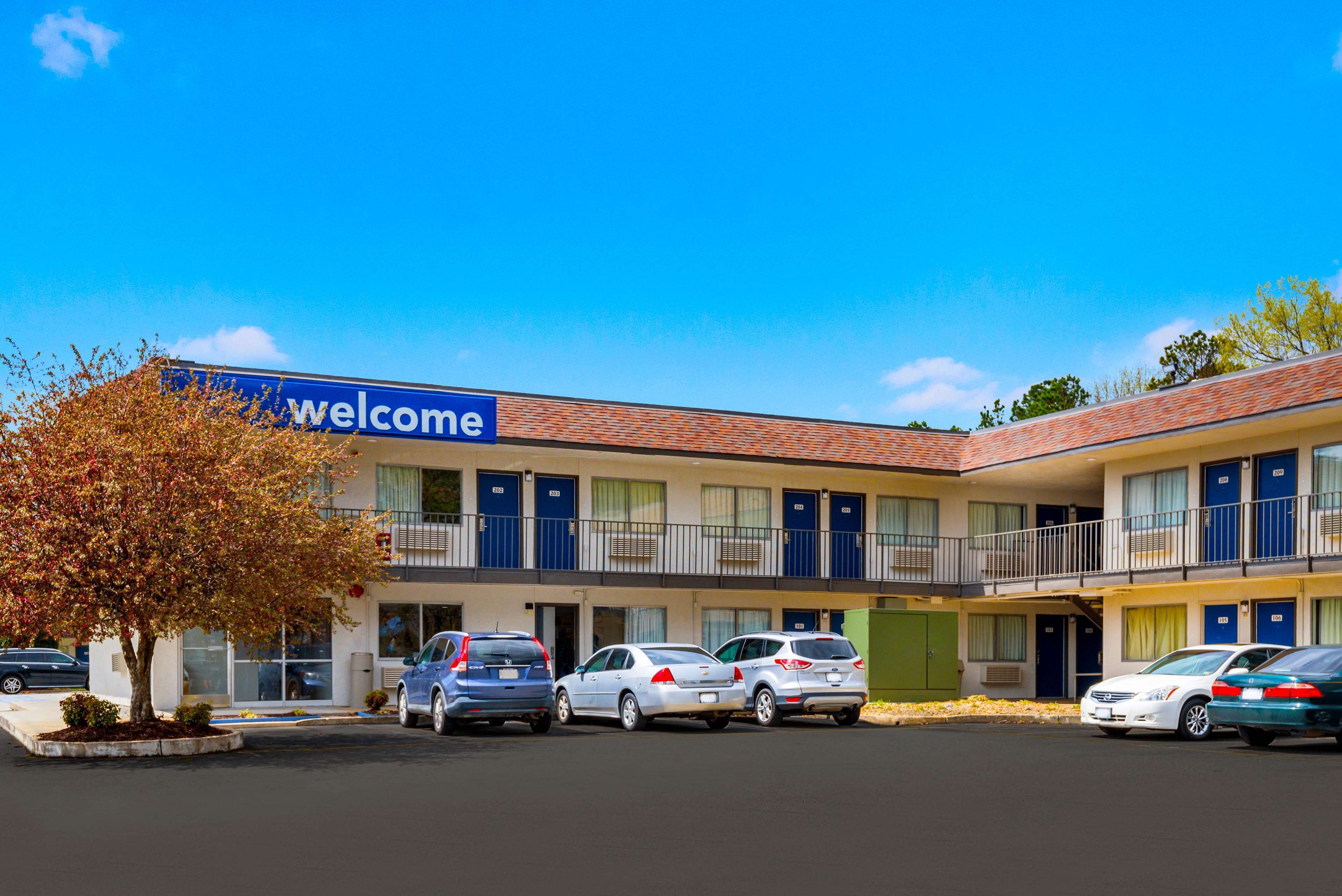 A motel with a 'Welcome' sign, multiple rooms, and a parking lot with several cars.