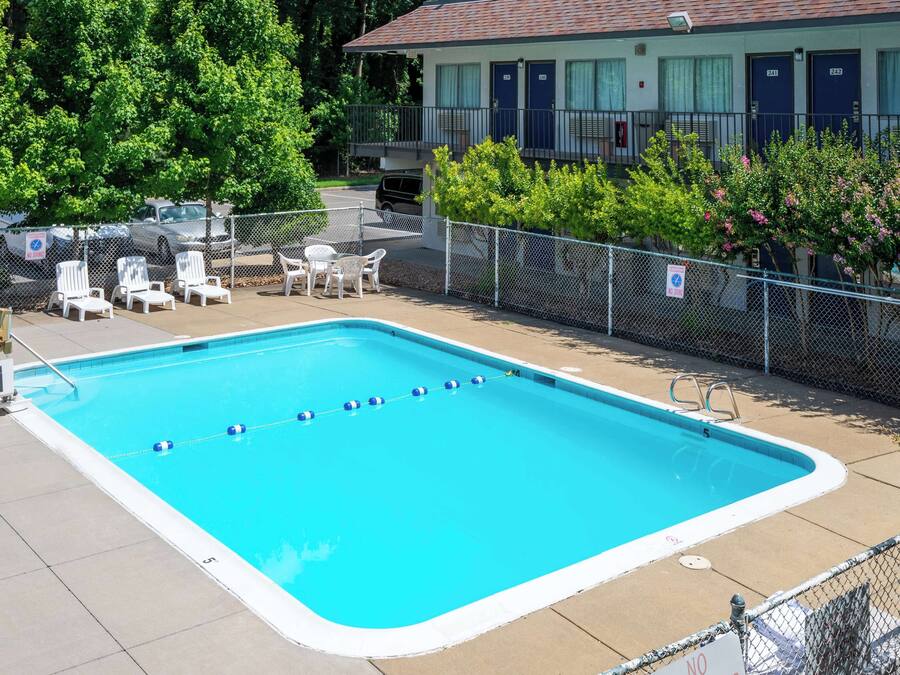 Une piscine rectangulaire bordée de blanc, entourée d’une clôture et jouxtant un bâtiment doté d’un balcon.