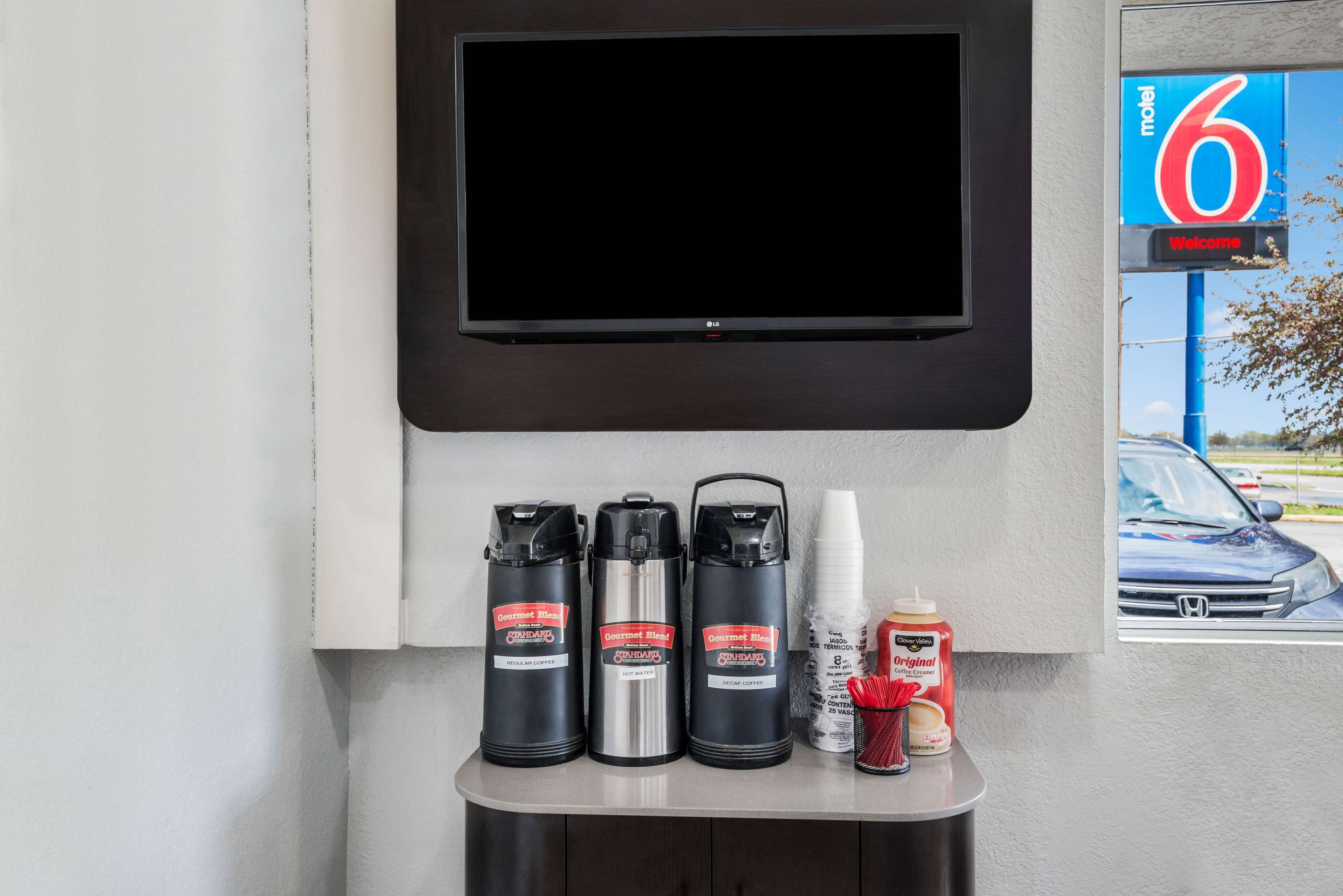 A wall-mounted TV above a counter with three coffee dispensers, a coffee cup, and a bottle of coffee creamer.