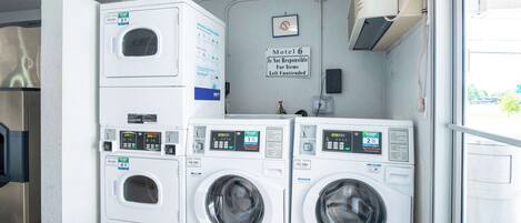 A laundry area with stacked washers and dryers, a sign indicating the motel's policy on unattended items, and a window with a view of greenery outside.