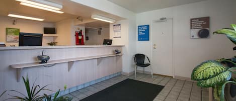A reception area with a counter, a computer, a fire extinguisher, a chair, and a plant.