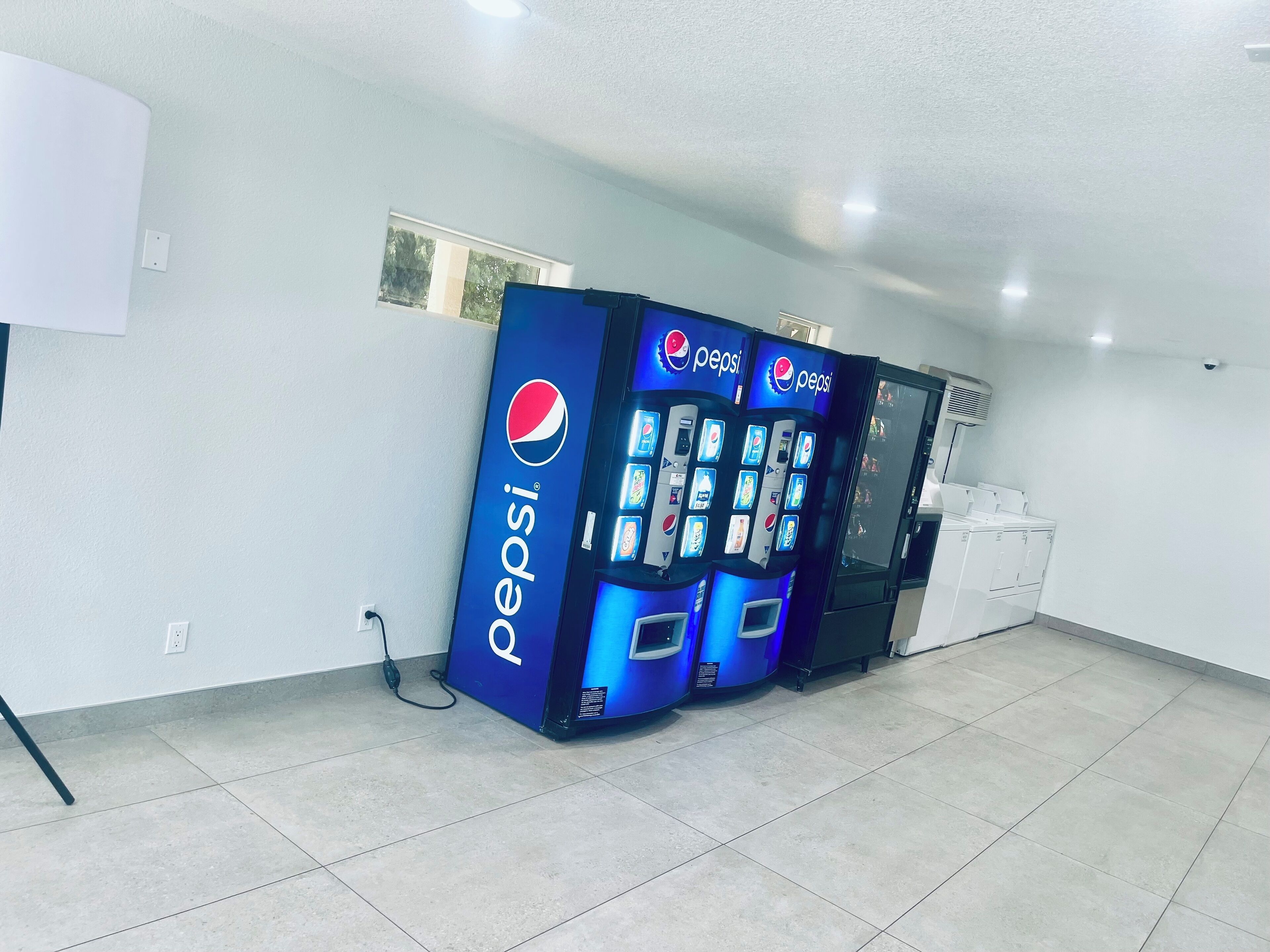 A room with a tiled floor, a white wall, and a row of Pepsi vending machines.