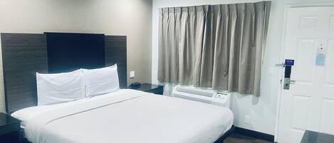 A hotel room with a bed, a bedside table, a wall-mounted air conditioner, and a door with a sign.