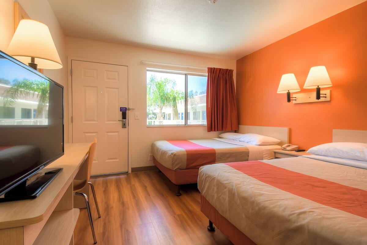 A hotel room with two beds, a desk with a TV, a window with a view of palm trees, and two wall-mounted lamps.