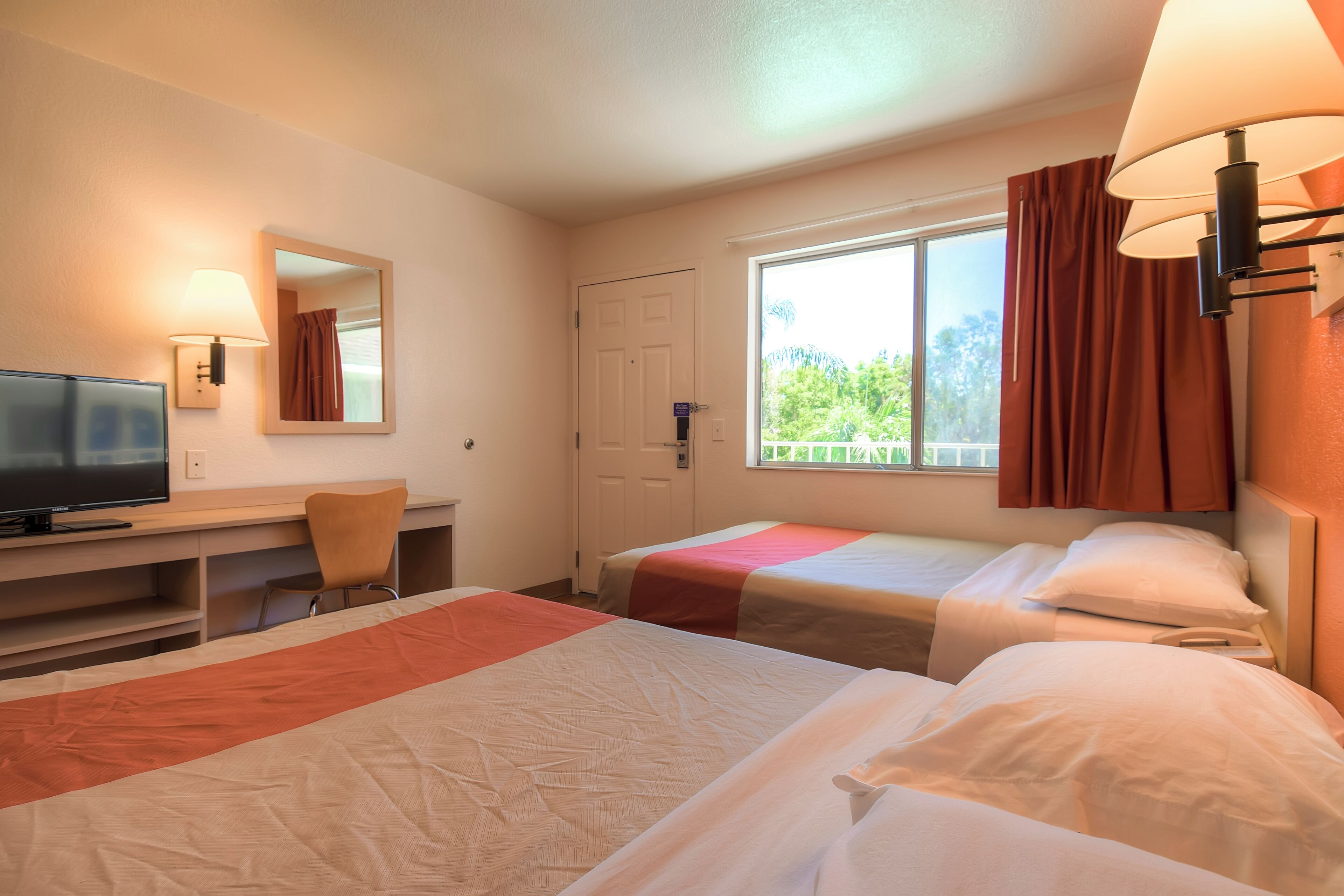 A hotel room with two beds, a desk with a television, a chair, and a window with curtains.