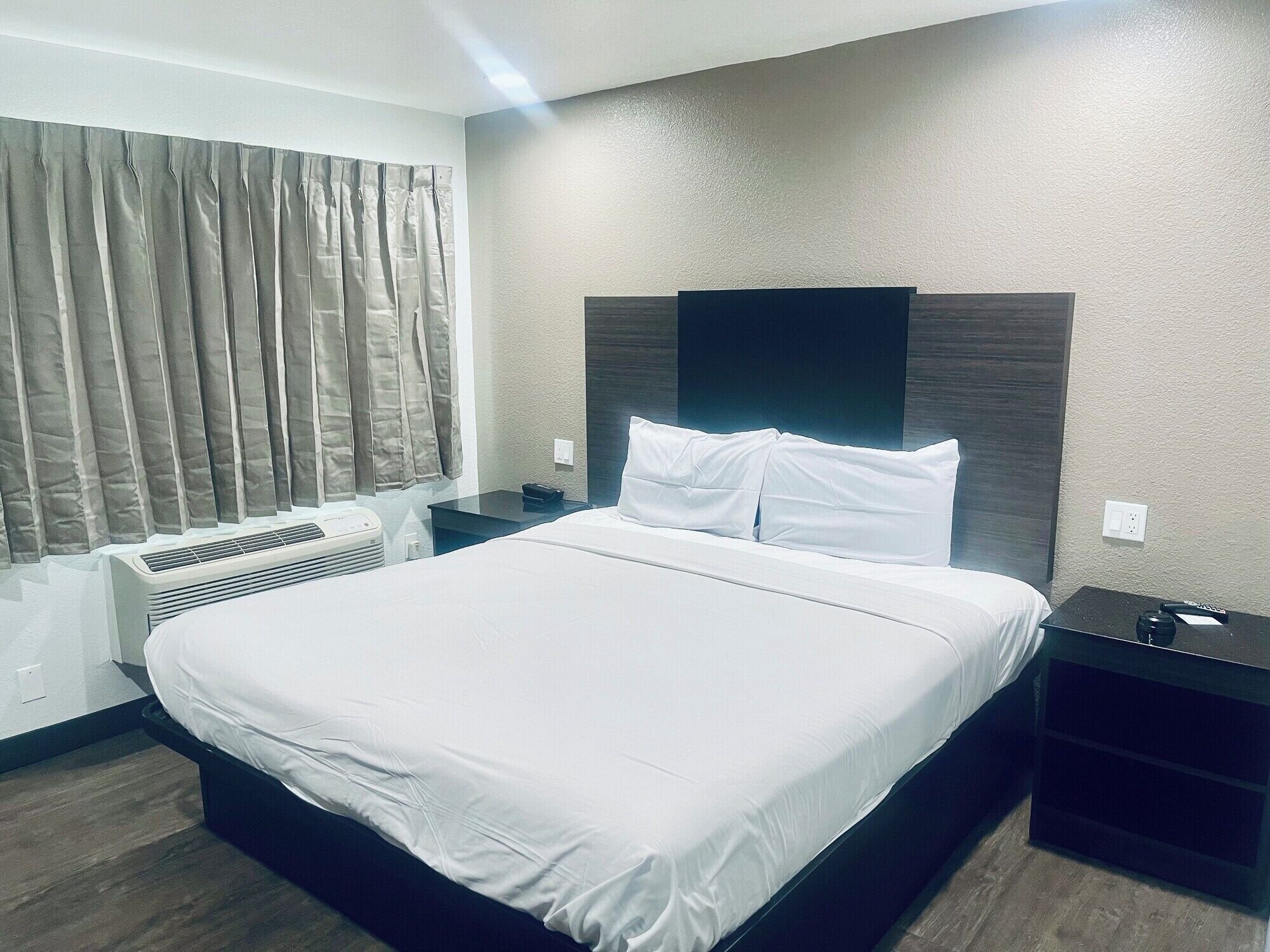 Una habitación de hotel con una cama grande, una mesita de noche, aire acondicionado instalado en la pared y una ventana con cortinas.