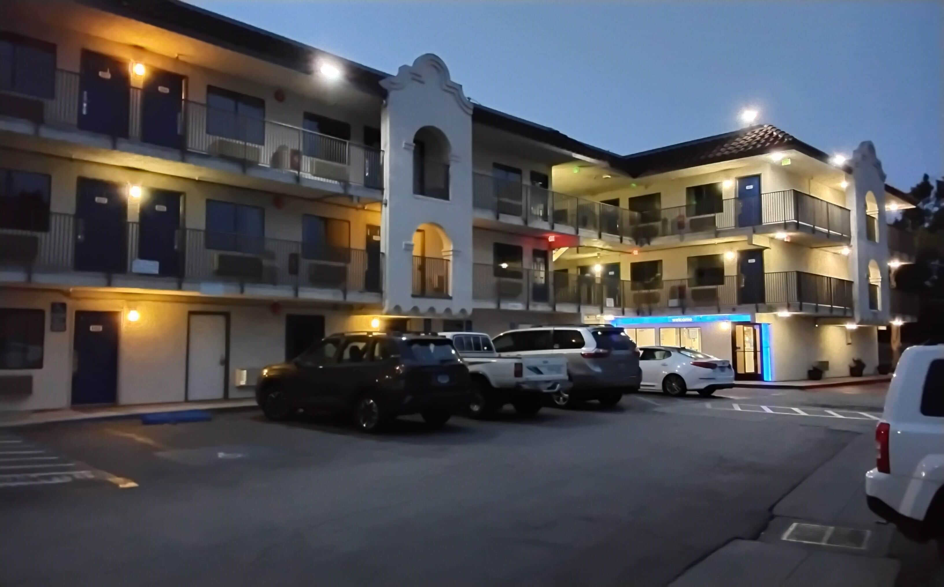 Un hotel de varios pisos con un patio central, zona de estacionamiento y un edificio con un cartel de 'Welcome'.
