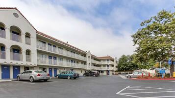 Un hotel de varios pisos con estacionamiento, que presenta una hilera de habitaciones con balcones, un cielo despejado y un árbol en primer plano.