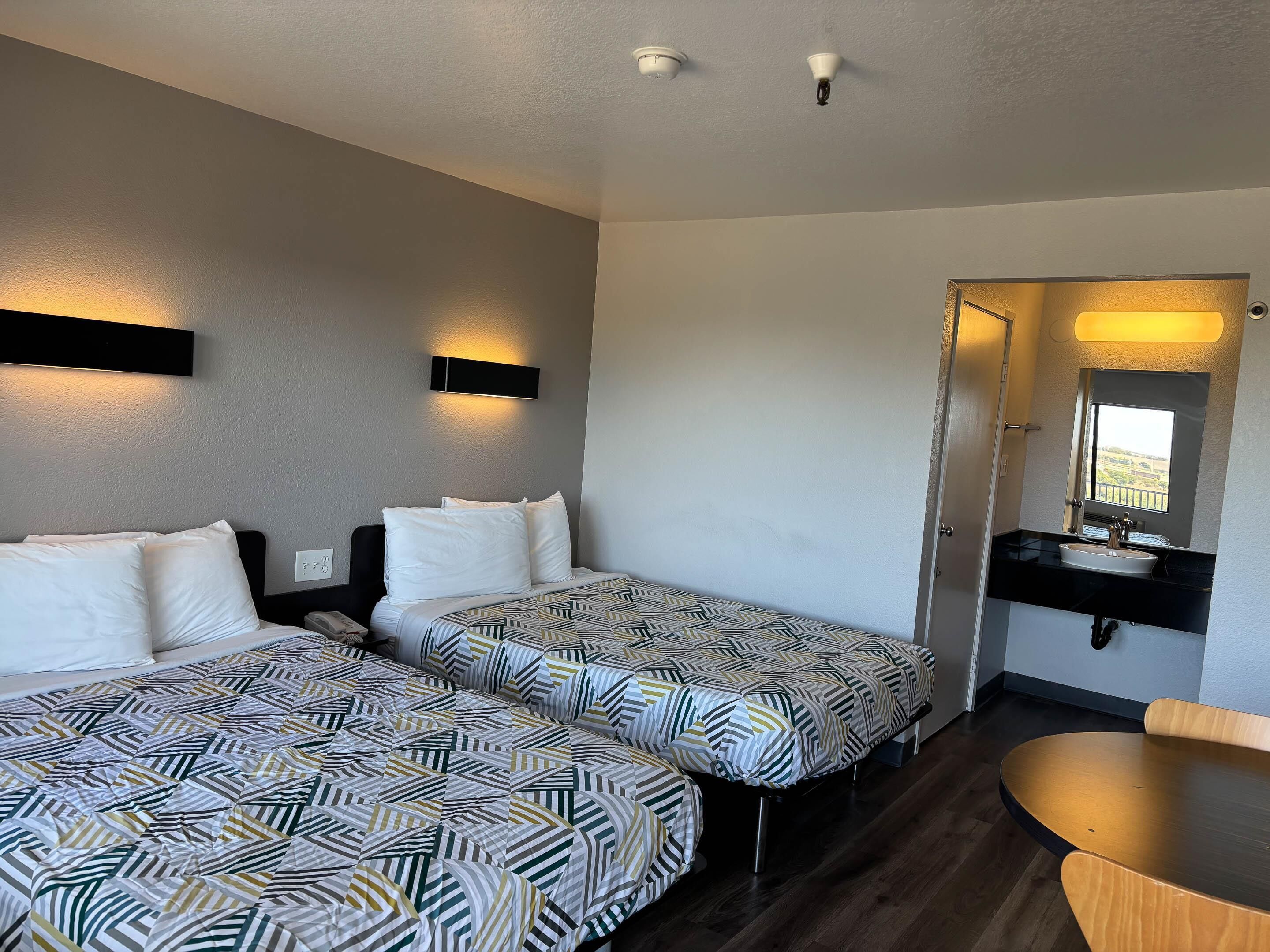Une chambre d’hôtel avec deux lits, une petite table, une salle de bain avec un miroir et une vue par la fenêtre.