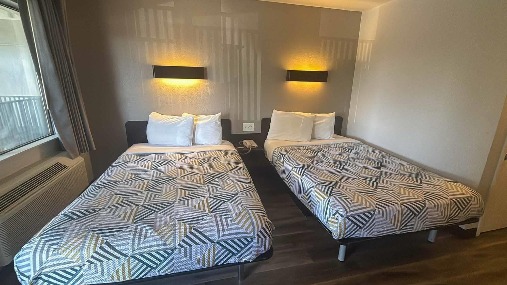 Two single beds with patterned bedding, a window with curtains, and wall-mounted lights.