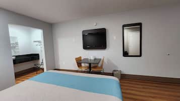A hotel room with a bed, a desk with two chairs, a flat-screen TV, a mirror, and a small trash bin.