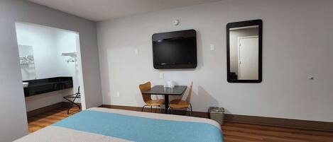 A hotel room with a bed, a desk with two chairs, a flat-screen TV, a mirror, and a small trash bin.