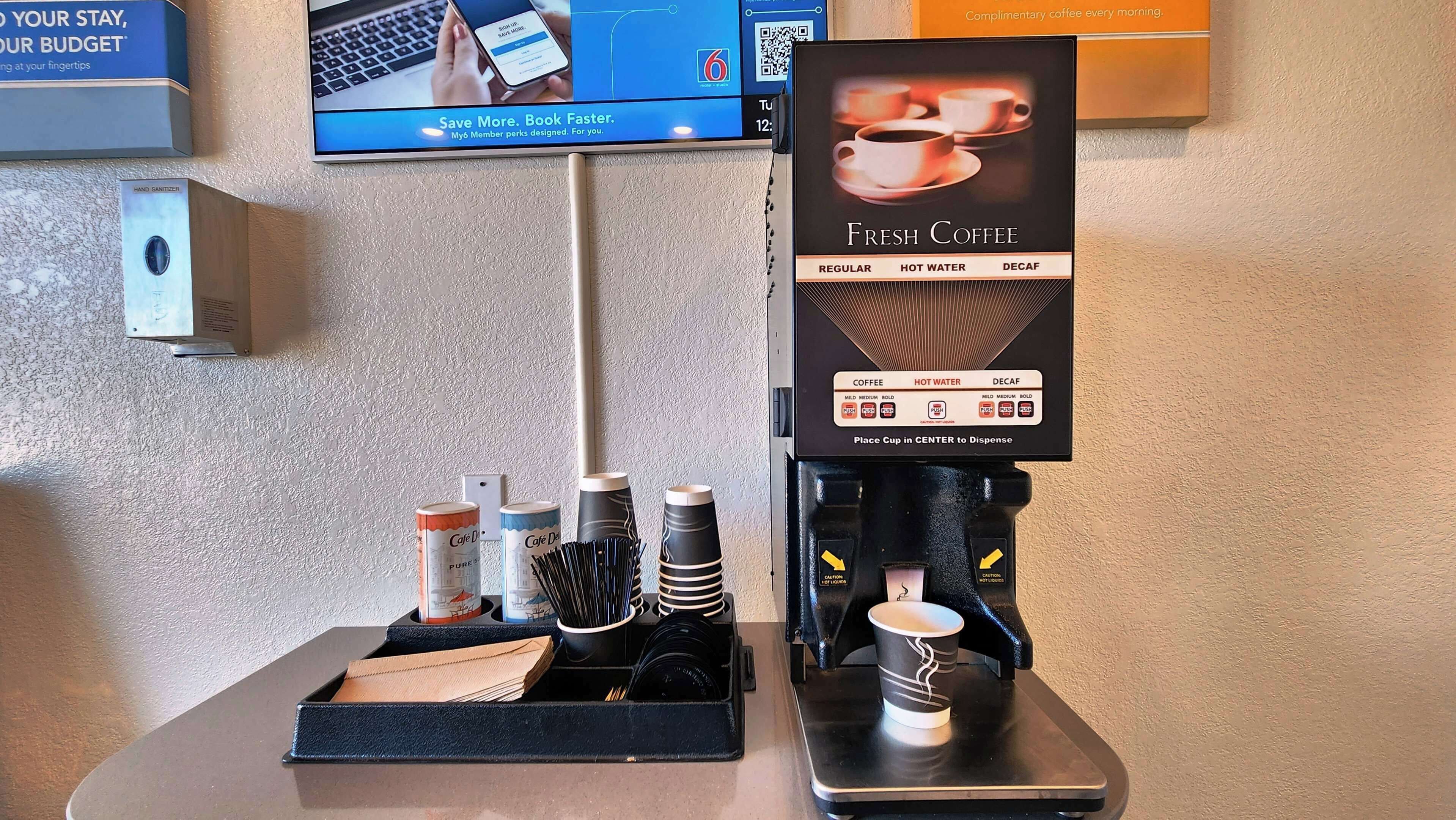 A coffee machine with a sign indicating it dispenses fresh coffee, hot water, and decaf. There are disposable cups and a hand sanitizer dispenser on the counter.