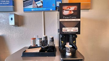 Máquina de café com aviso indicando que fornece café fresco, água quente e café descafeinado. Há copos descartáveis e um dispensador de álcool em gel no balcão.