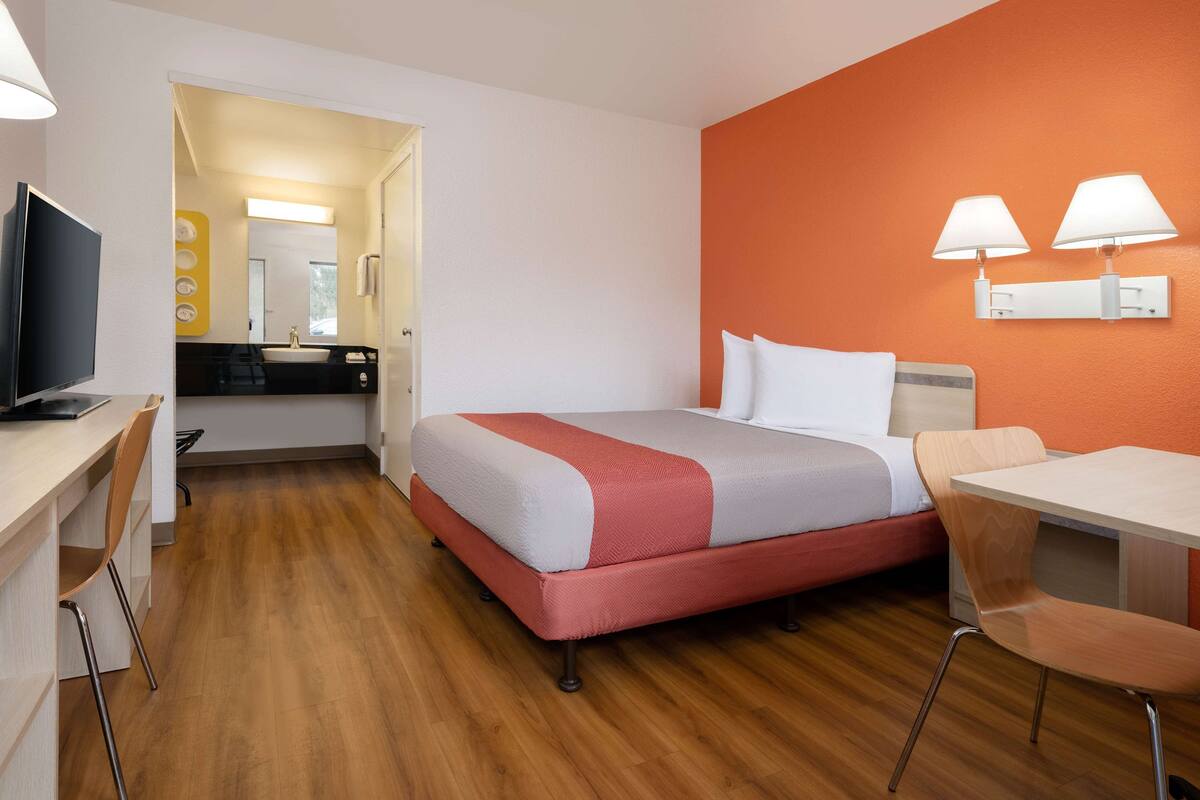 A hotel room with a bed, a desk with a TV, a chair, a table, and a bathroom visible through an open door.