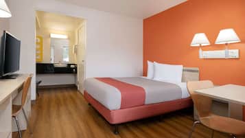 A hotel room with a bed, a desk with a TV, a chair, a table, and a bathroom visible through an open door.