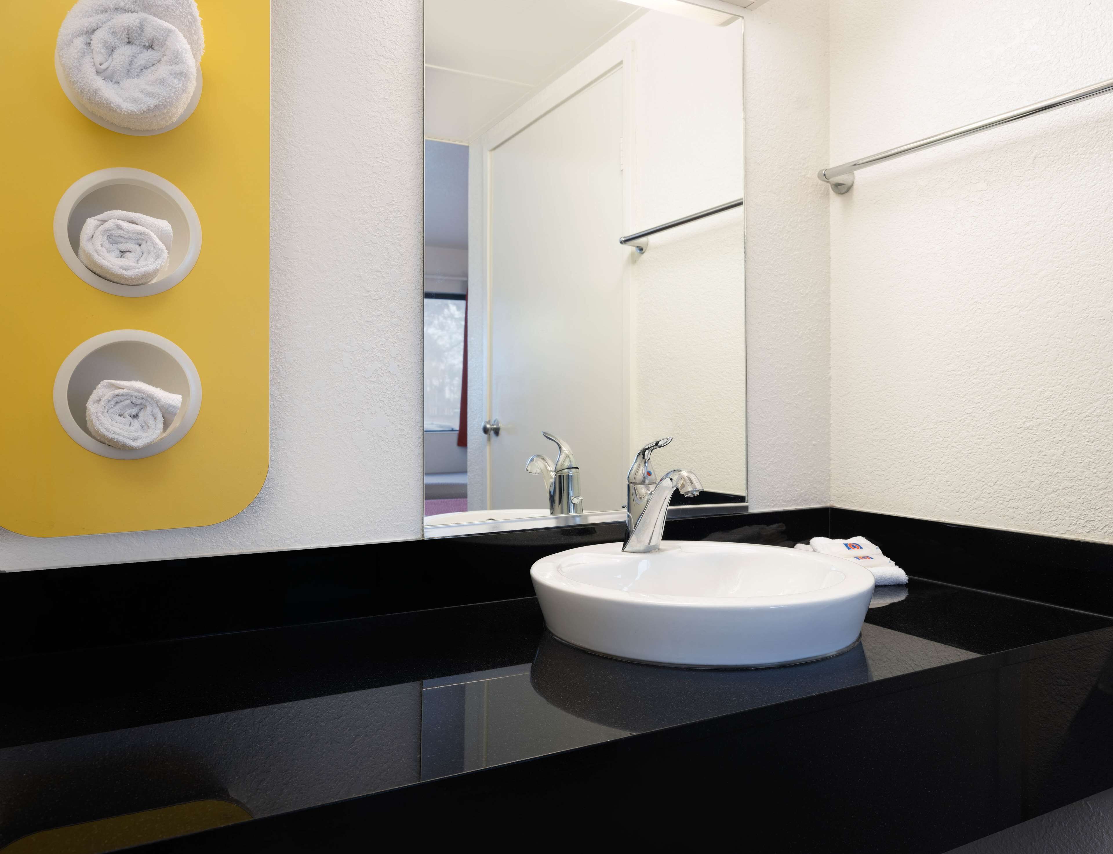 Un baño moderno con encimera negra, lavabo blanco y tres toallas blancas sobre una pared amarilla.