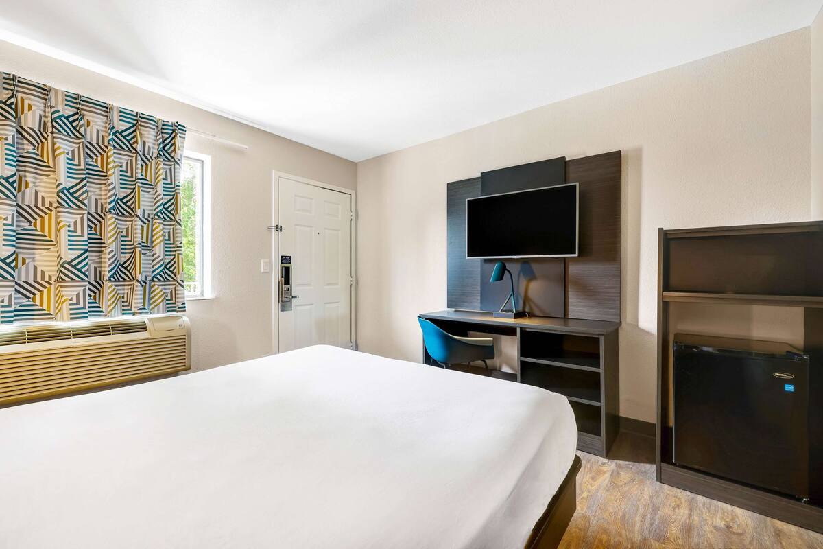Een hotelkamer met een bed, een bureau met stoel, een flatscreen televisie, een minibar en een raam met gordijnen met een patroon.