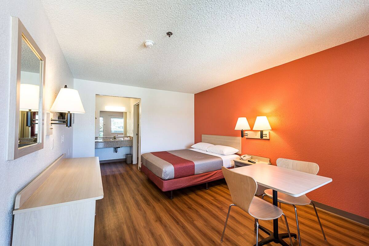 A hotel room with a bed, a desk, a chair, and a table. There is a mirror on the wall and a lamp on the desk.