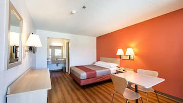 Habitación de hotel con cama, escritorio, silla y mesa. Hay un espejo en la pared y una lámpara sobre el escritorio.