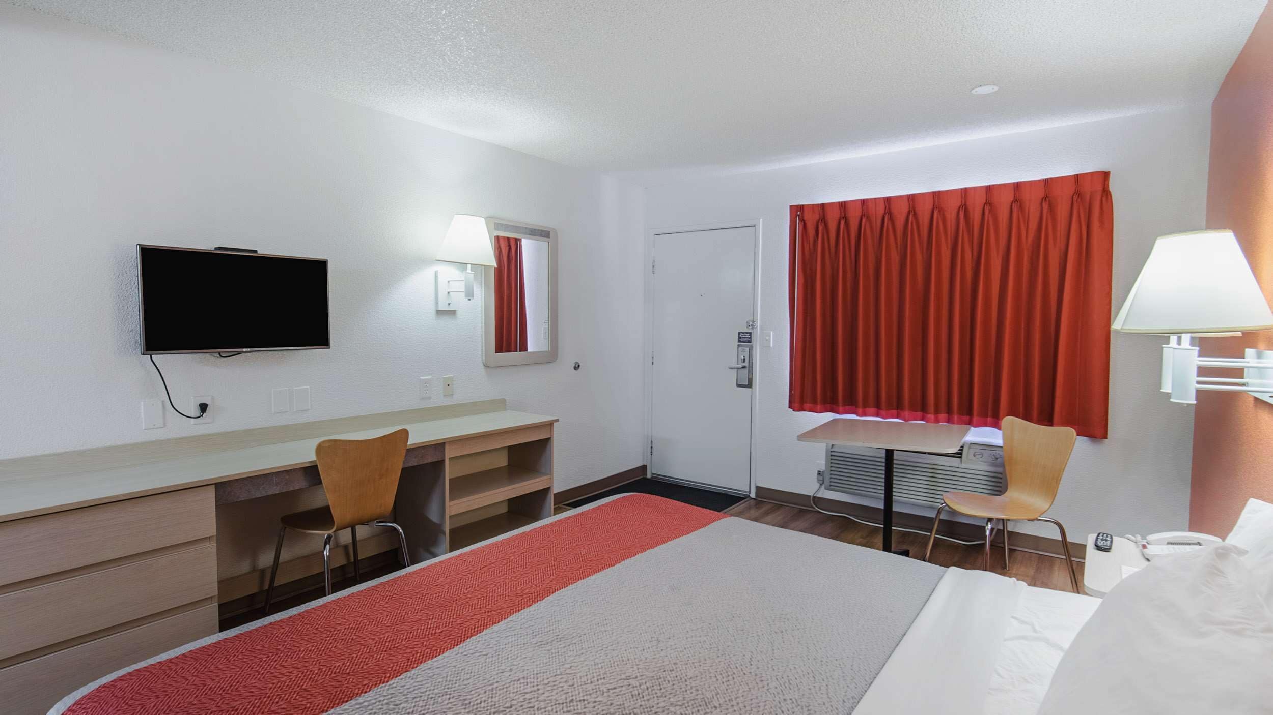A hotel room with a bed, a desk, two chairs, a television, a window with red curtains, and a wall-mounted lamp.