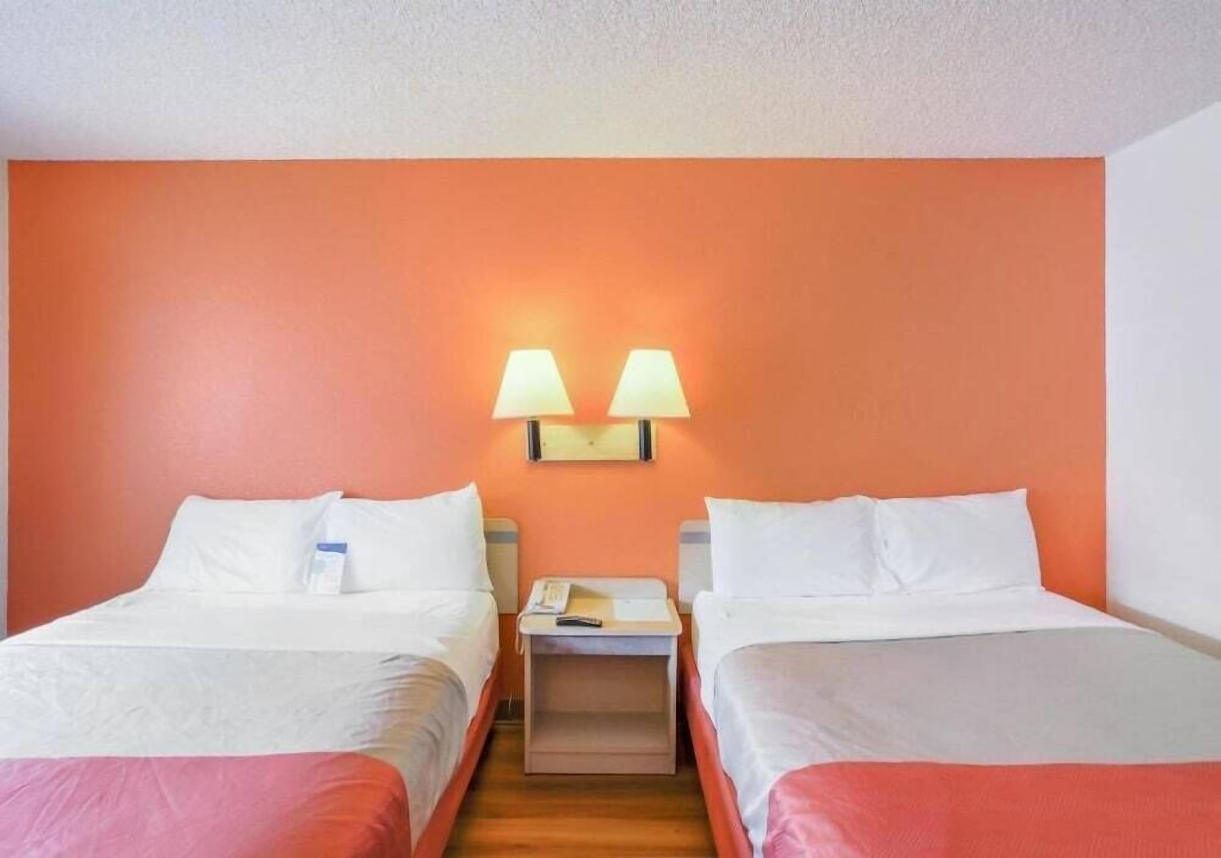 A hotel room with two beds, a nightstand with a lamp, and a wall-mounted light fixture.