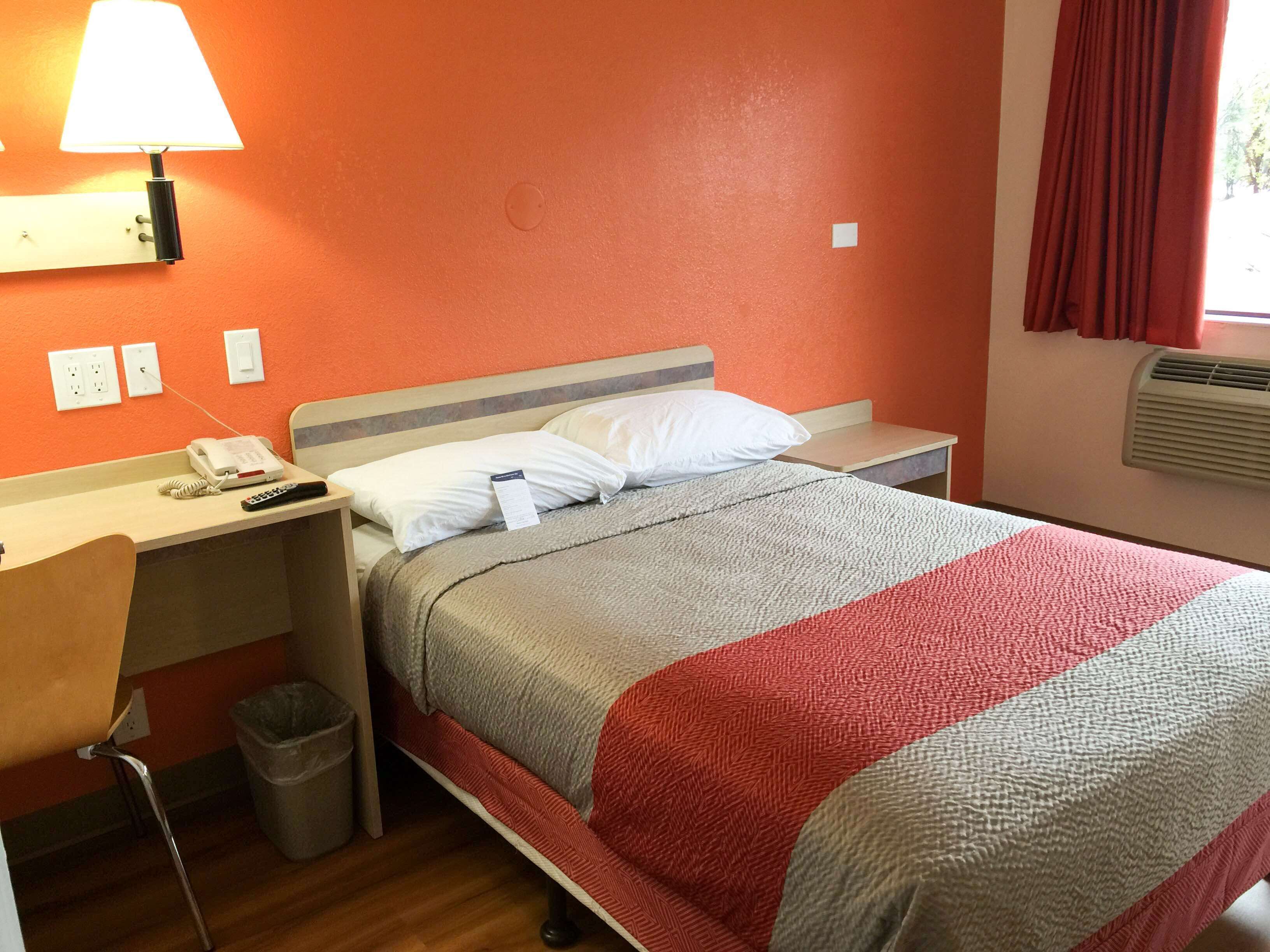 A hotel room with a bed, a nightstand with a lamp, a chair, a trash bin, and a window with curtains.