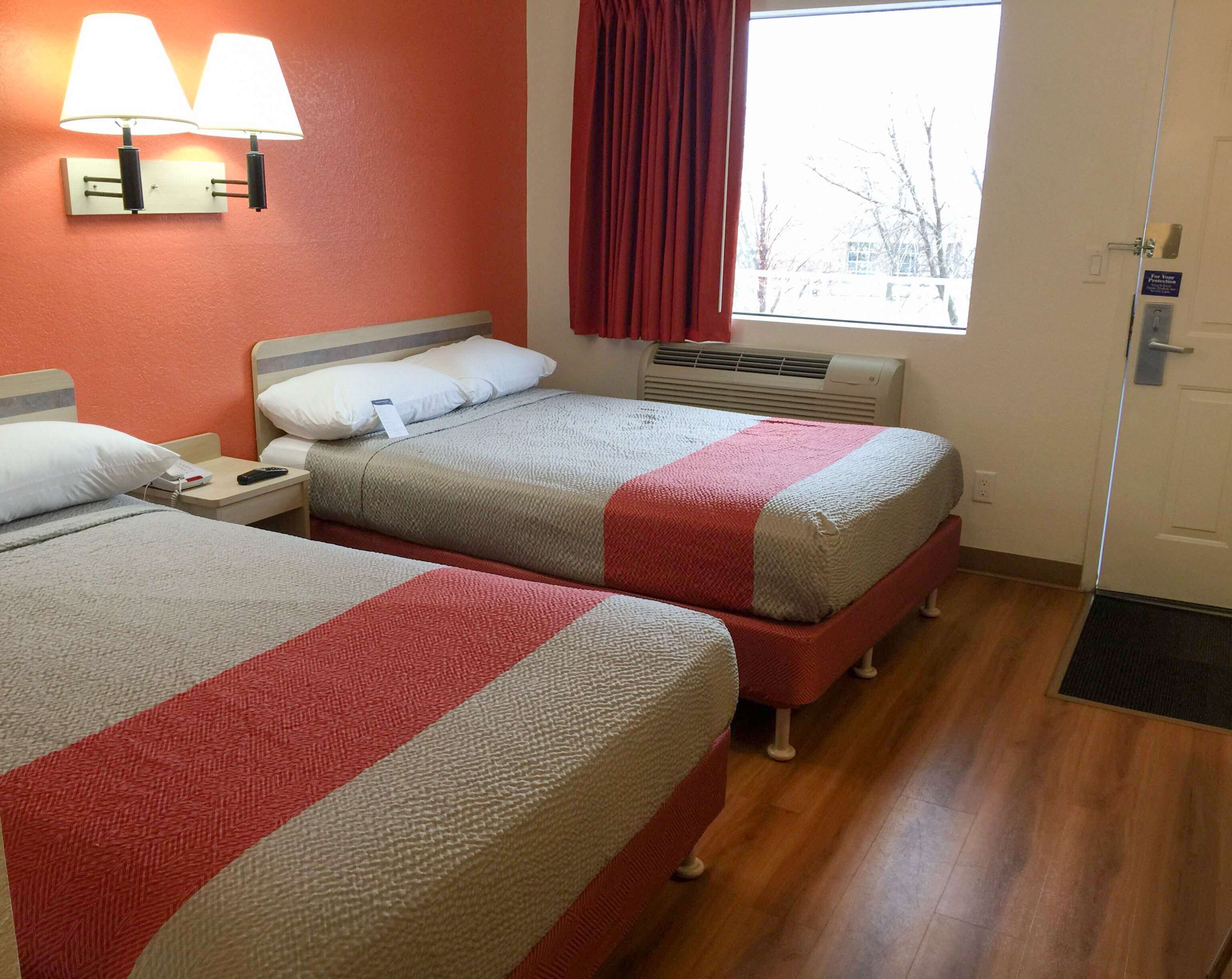 Two single beds with red and white bedding, a wall-mounted lamp, a window with a red curtain, and a door with a sign.