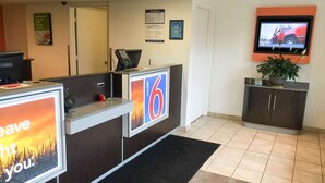 Een receptiebalie met een digitaal scherm, een aan de muur bevestigde televisie en een kamerplant.