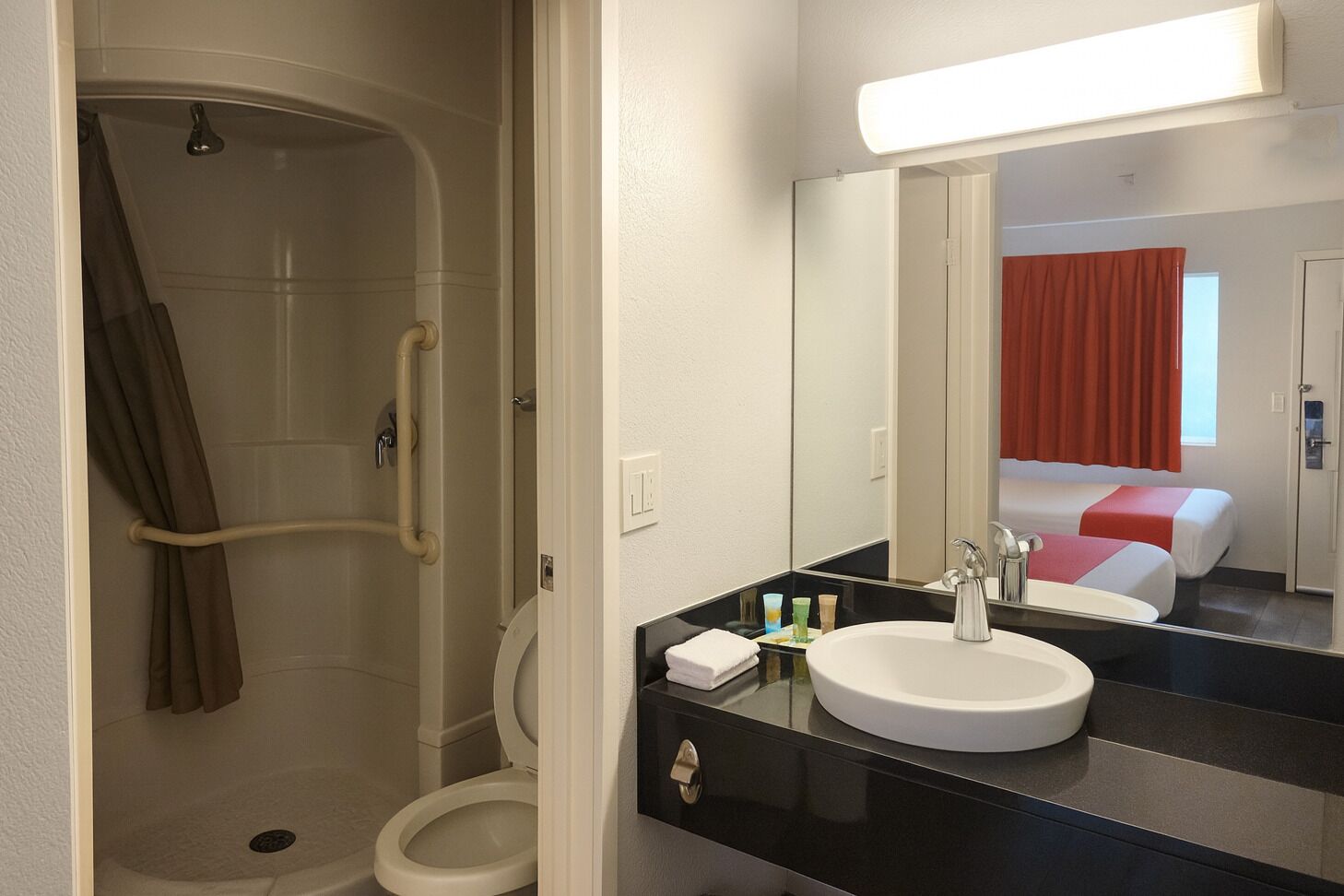 A bathroom with a shower, toilet, and sink. There is a bed visible in the background.