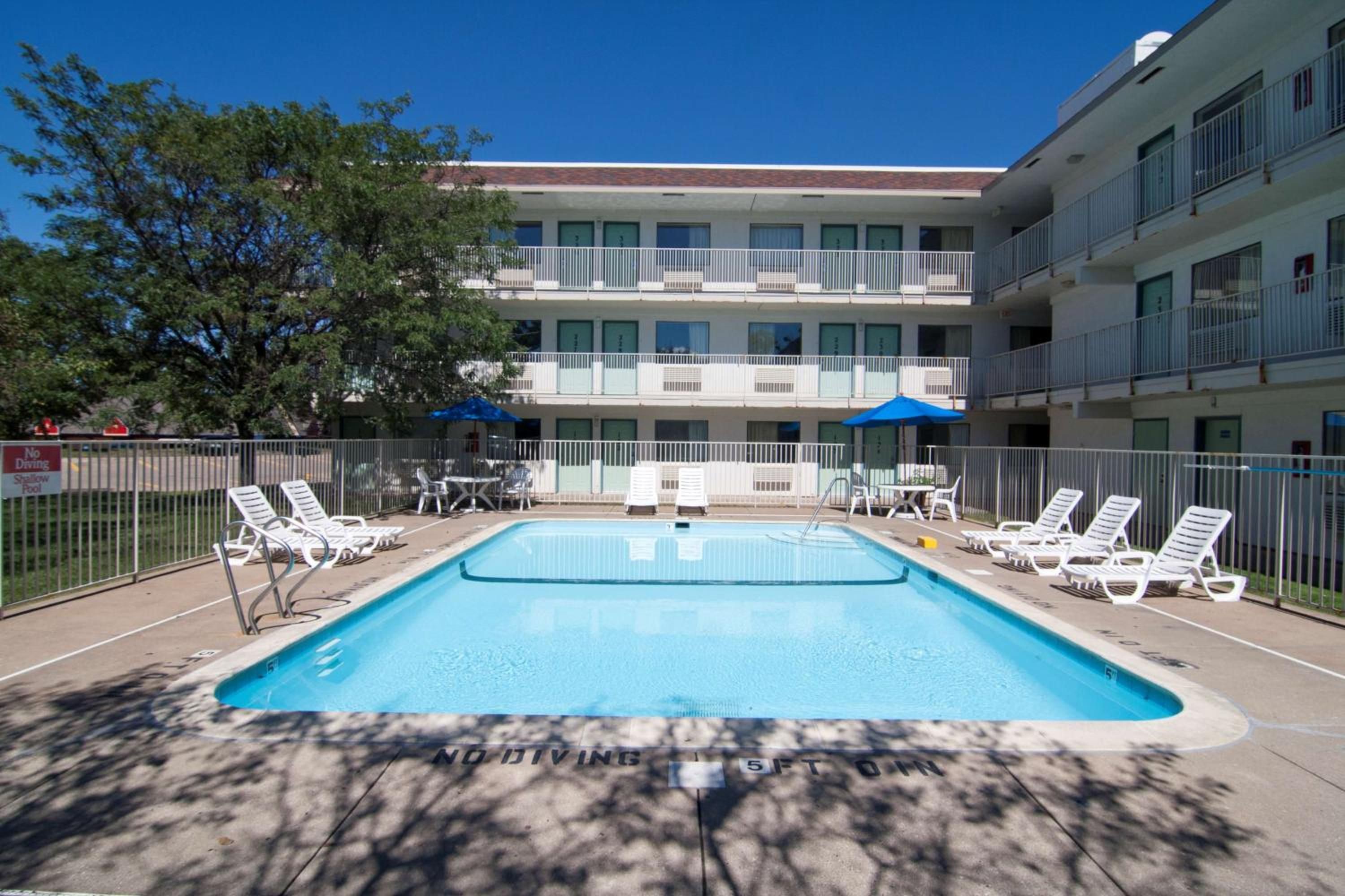A swimming pool with lounge chairs and umbrellas, surrounded by a fence, with a multi-story building in the background.