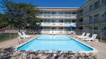 A swimming pool with lounge chairs and umbrellas, surrounded by a fence, with a multi-story building in the background.