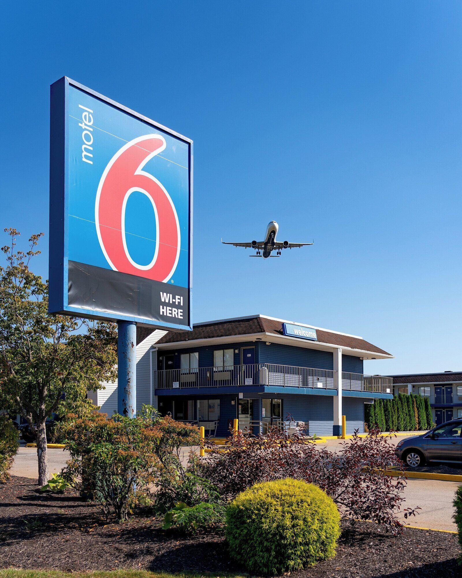 Letrero de motel con un '6' grande y el texto 'Wi-Fi aquí', un edificio con un letrero de 'Bienvenido' y un avión volando al fondo.