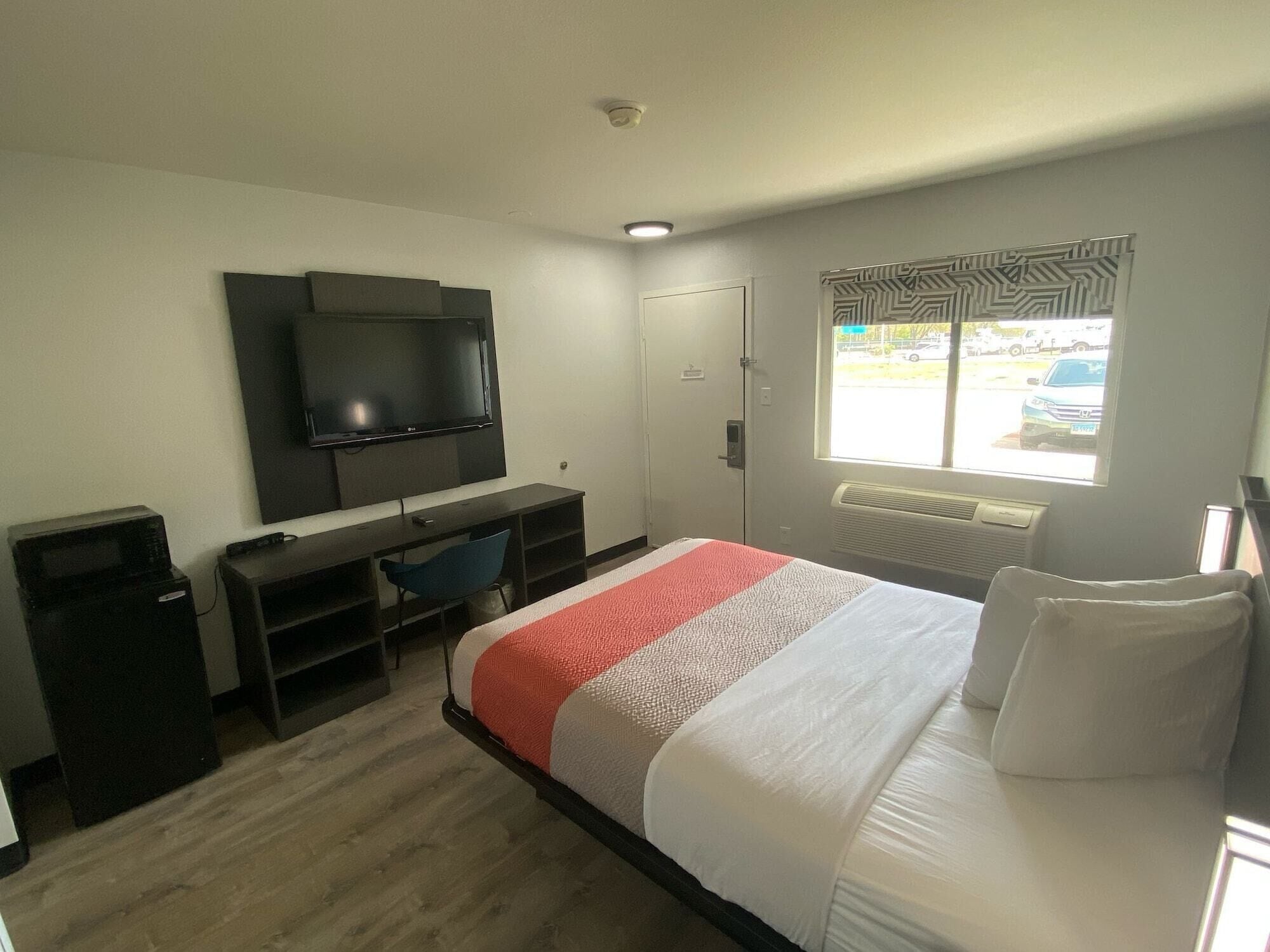 A hotel room with a bed, a desk with a chair, a television, a window with a view of the outside, and a radiator.