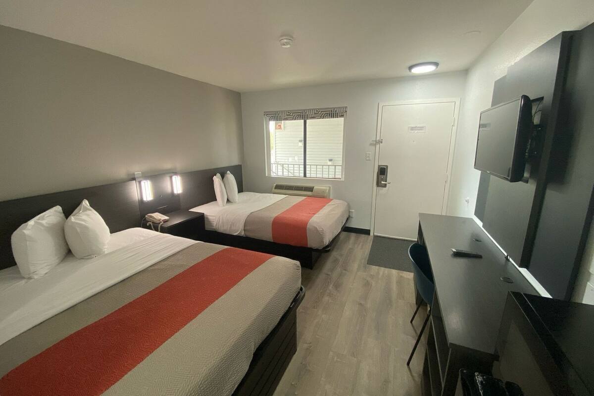 A hotel room with two beds, a desk, a TV, and a window with blinds.