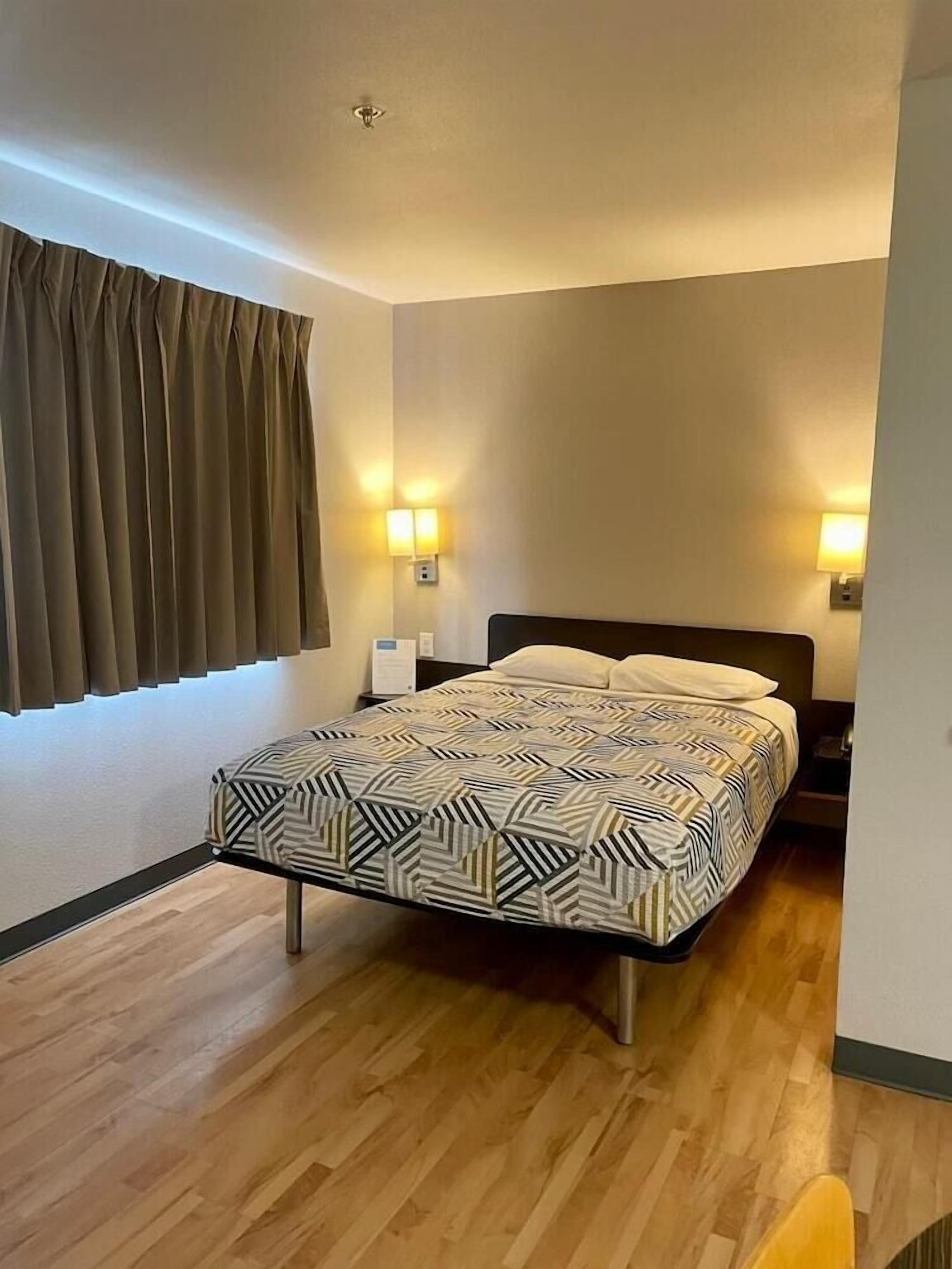 Un dormitorio con cama, piso de madera, ventana con cortinas e iluminación en la pared.