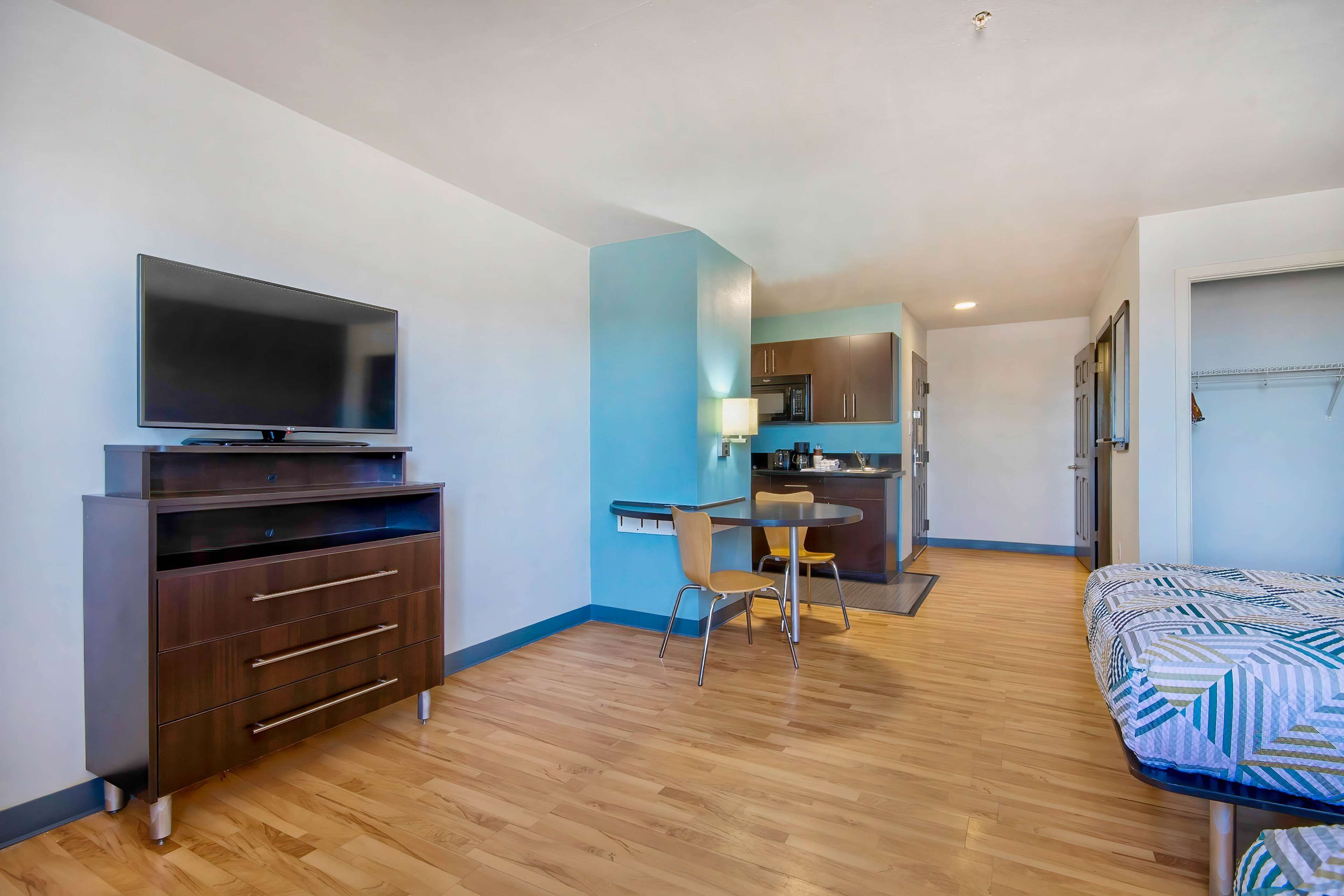 A compact hotel room with a flat-screen TV, a dark wooden dresser, a small dining table with two chairs, and a bed with a patterned bedspread.