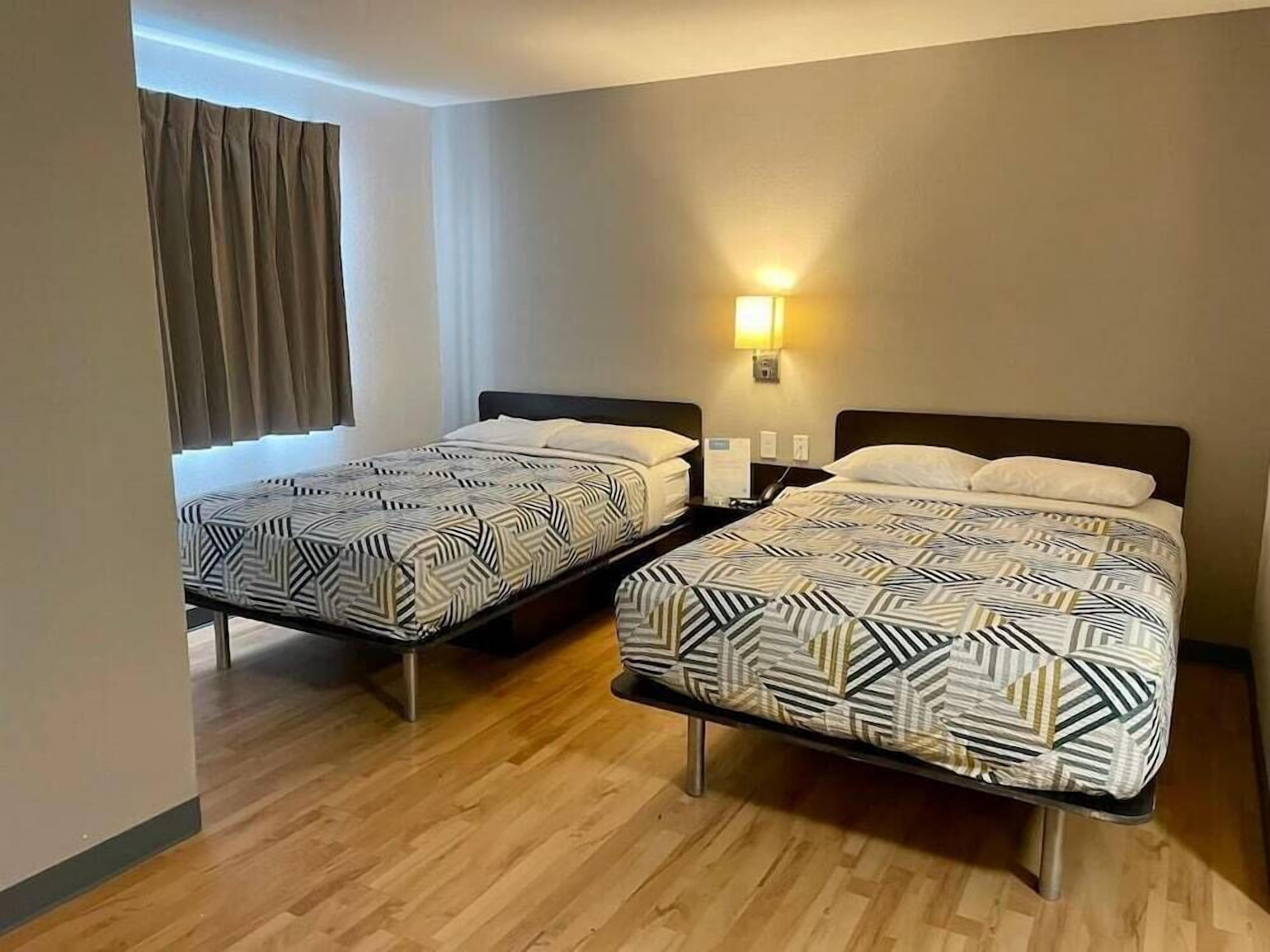 Ein Hotelzimmer mit zwei Betten, Holzboden, einem fenster mit Vorhängen und einer Wandleuchte.
