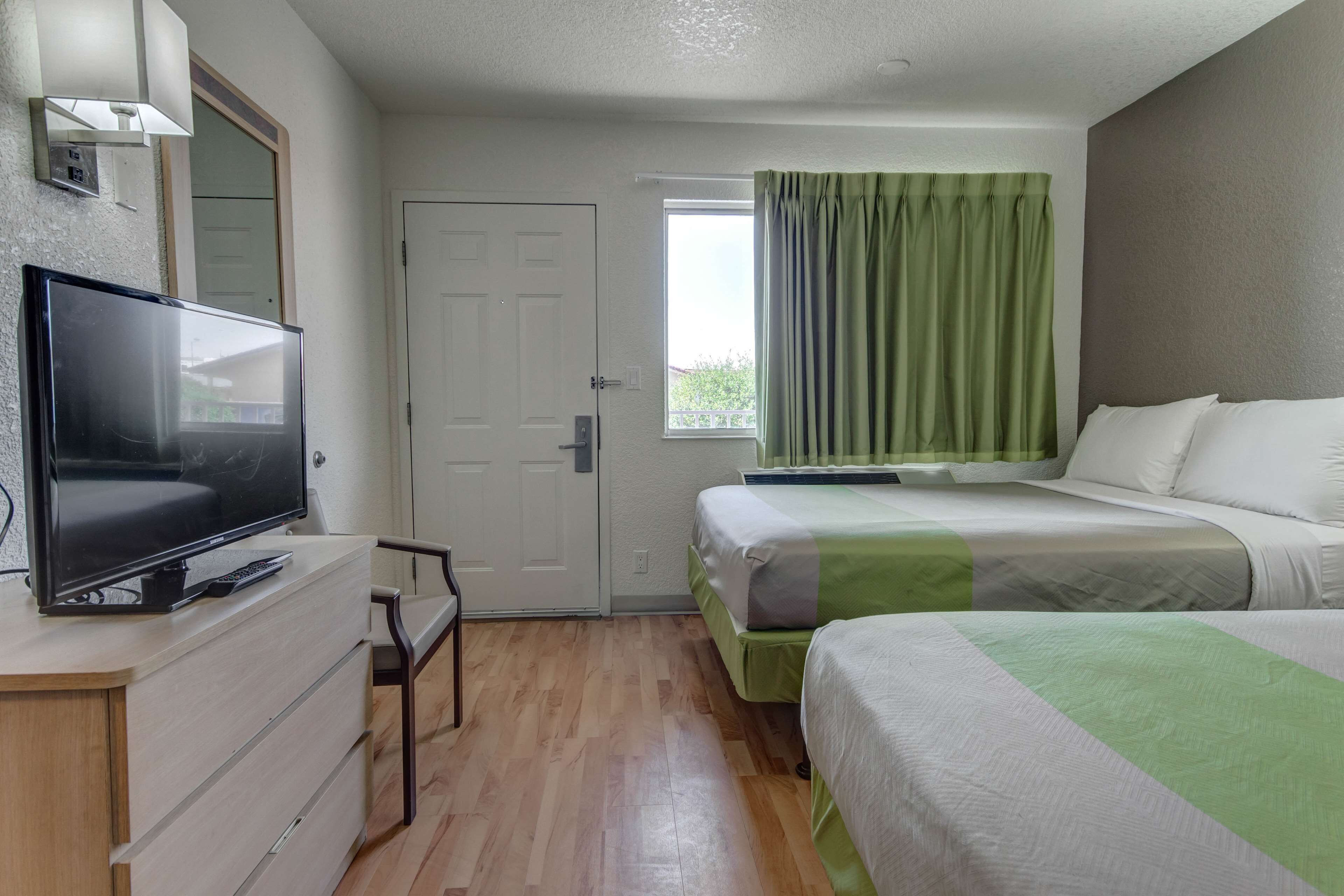 A hotel room with a flat-screen TV on a dresser, a door, a window with green curtains, and two beds with white and green bedding.