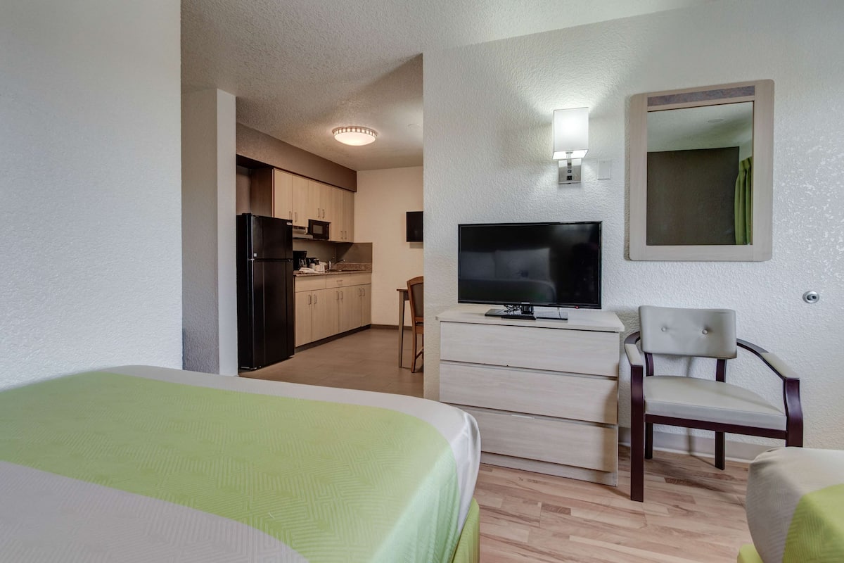 Quarto de hotel com cama, televisão, secretária e cadeira. Cozinha compacta ao fundo.