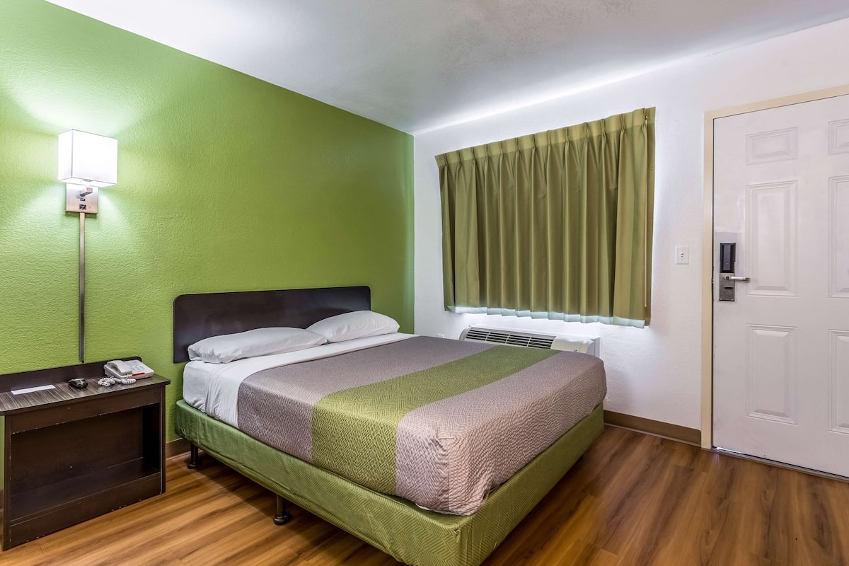 Quarto de hotel com parede verde, cama com manta verde e rosa, mesa de cabeceira com telefone e luminária, janela com cortinas e uma porta.