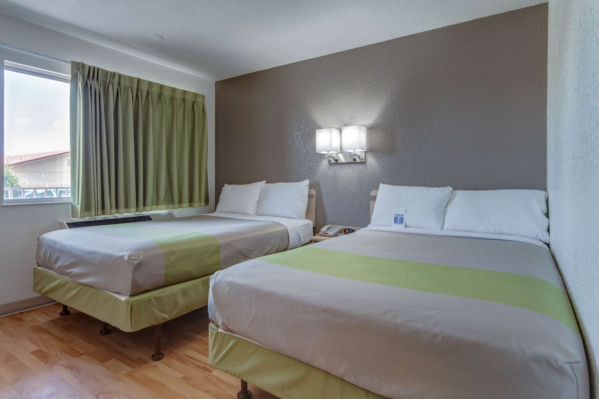 Quarto de hotel com duas camas, uma janela com cortinas, luminária fixada na parede e piso de madeira.