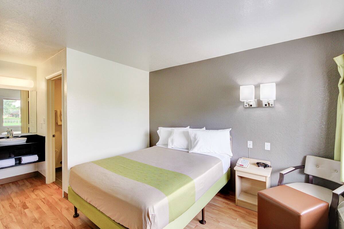 A hotel room with a bed, a bedside table with a lamp, a chair, and a bathroom with a sink and mirror.